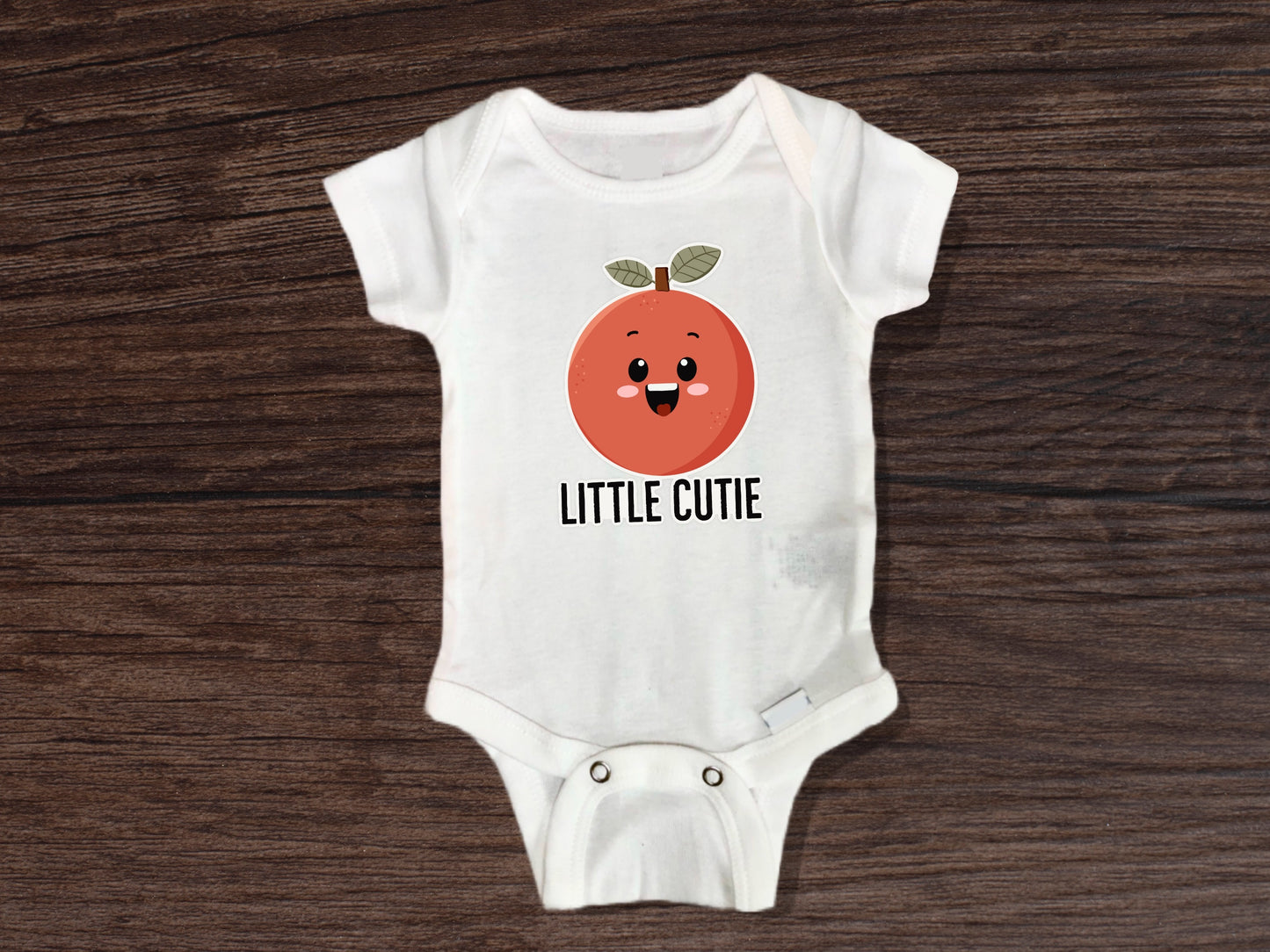 Adorable baby onesie with a cute orange fruit design and the phrase “Little Cutie” printed on the front; white short sleeve cotton bodysuit displayed flat on a wooden surface, surrounded by fresh whole clementines and green leaves for a vibrant citrus-themed look; perfect baby shower gift, newborn outfit, or fun baby clothing for fruit lovers; soft, breathable fabric ideal for infants and easy snap closures for quick diaper changes. Great choice for citrus themed baby shower.