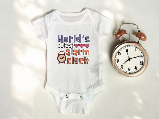 Adorable white baby onesie with original design that says “World’s Cutest Alarm Clock” in colorful, playful fonts, accented with three pink hearts and a cute cartoon alarm clock illustration. Perfect for parents shopping funny baby clothes, sleep-themed infant outfits, or unique newborn baby shower gifts. The design is sublimated onto the fabric using printable vinyl for a vibrant and lasting finish. Created in-house by the shop owner. This one-of-a-kind handmade design adds a humorous touch to your baby’s 