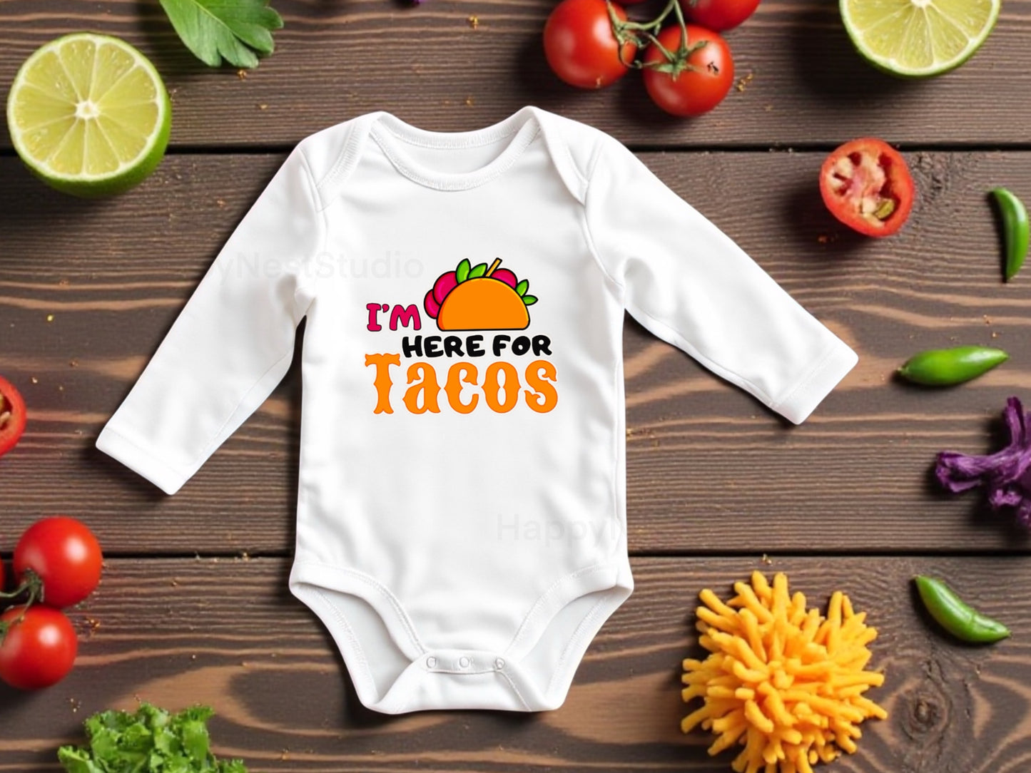 White long sleeve baby onesie with 'I'm here for tacos' text on a wooden surface with vegetables.